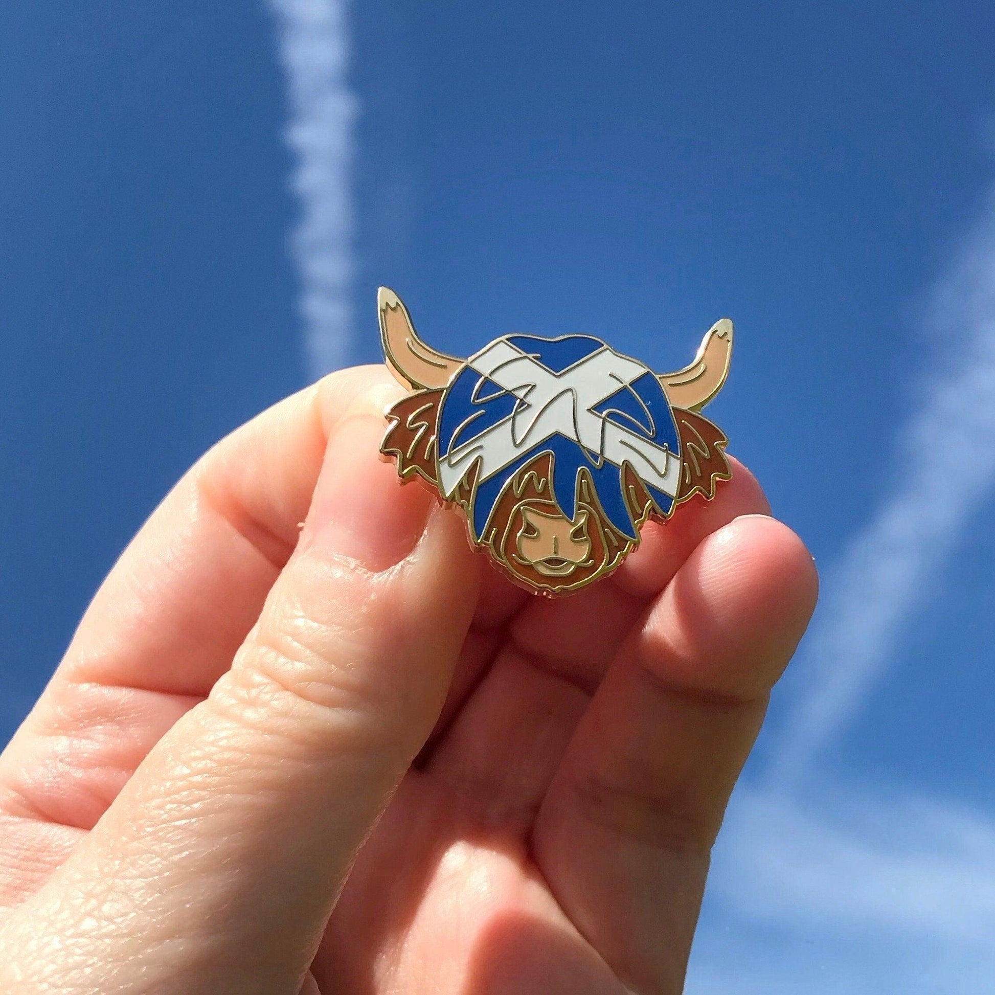 A photo of a blue sky with a Highland Cow Enamel Pin with a blue and white Saltire flag in its hair