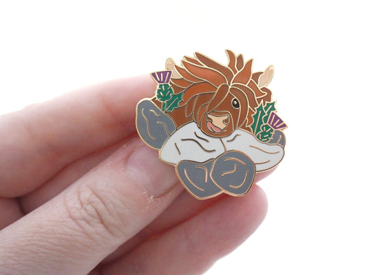 A photo of a funny highland cow enamel pin with thistles and a wall